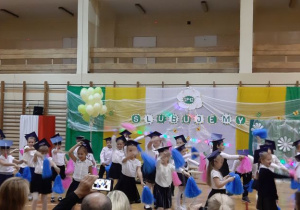 Ślubowanie I klas- pierwszaki w czapeczkach z pomponami tańczą podczas uroczystości na sali gimnastycznej.