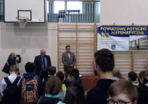 na ścianie sali gimnastycznej duży napis powiatowe potyczki matematyczne, przed zebranymi uczniami dwóch mężczyzn, jeden z nich mówi do mikrofonu