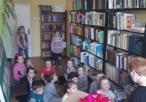 Uczniowie klasy 1 w bibliotece słuchają bajki czytanej przez panią bibliotekarkę.