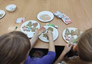 Dzieci siedzą przy stolikach i ozdabiają świąteczne pierniki.