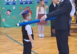 Pan dyrektor pasuje uczniów z klas pierwszych - dużym ołówkiem dotyka ramienia chłopca..