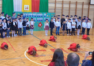 Na tle dzieci z klas pierwszych, na sali gimnastycznej uczniowie w czerwonych strojach wykonują układ taneczny,